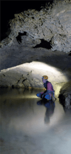 La photographie montre une cavité souterraine horizontale. Une personne y est accroupie et de l’eau lui recouvre les pieds.